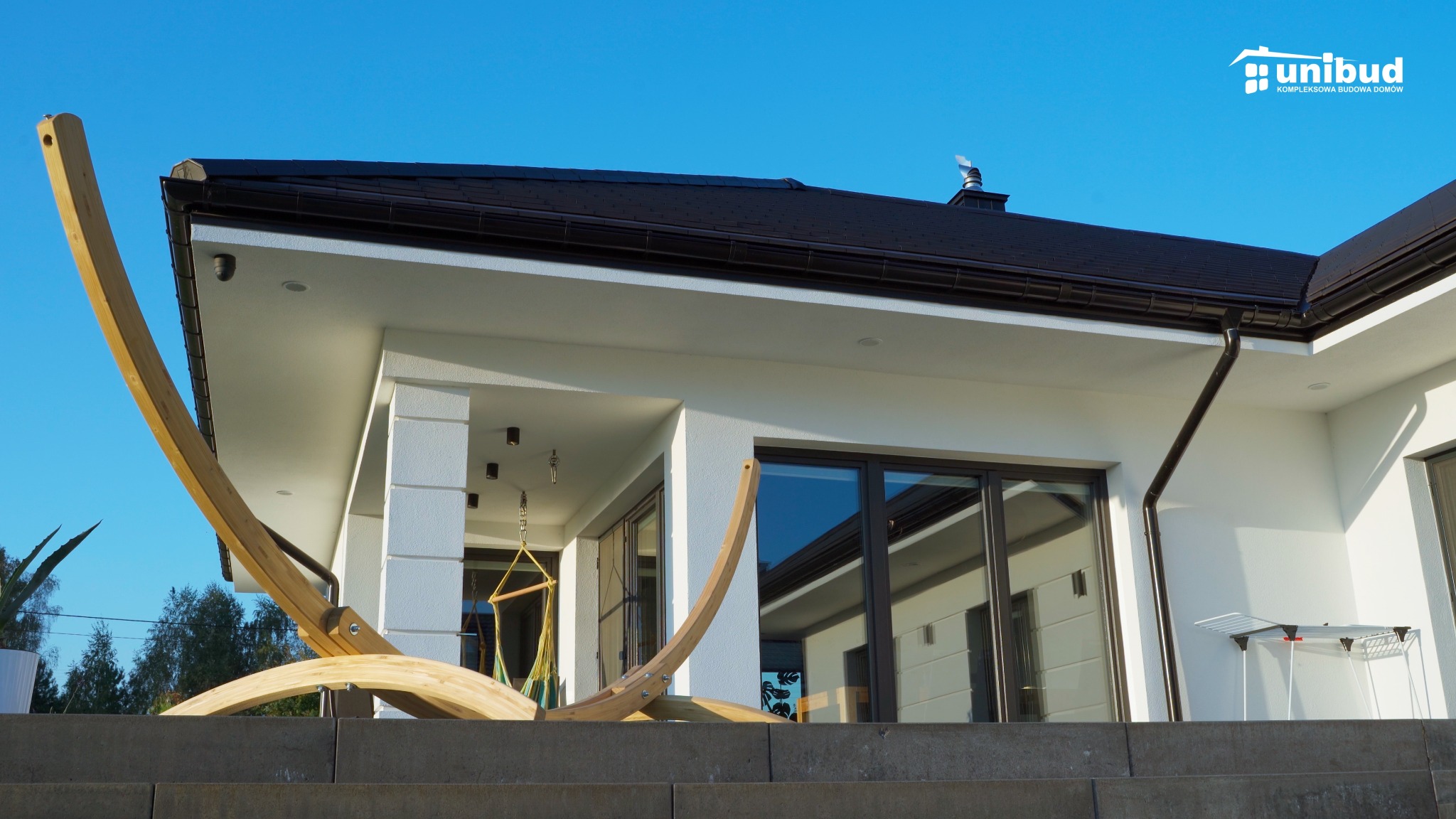 Nowoczesny dom jednorodzinny z białymi ścianami, ciemnobrązowym dachem i drewnianą konstrukcją huśtawki hamakowej na pierwszym planie. Widoczne okna z refleksami i elementy wykończeniowe.