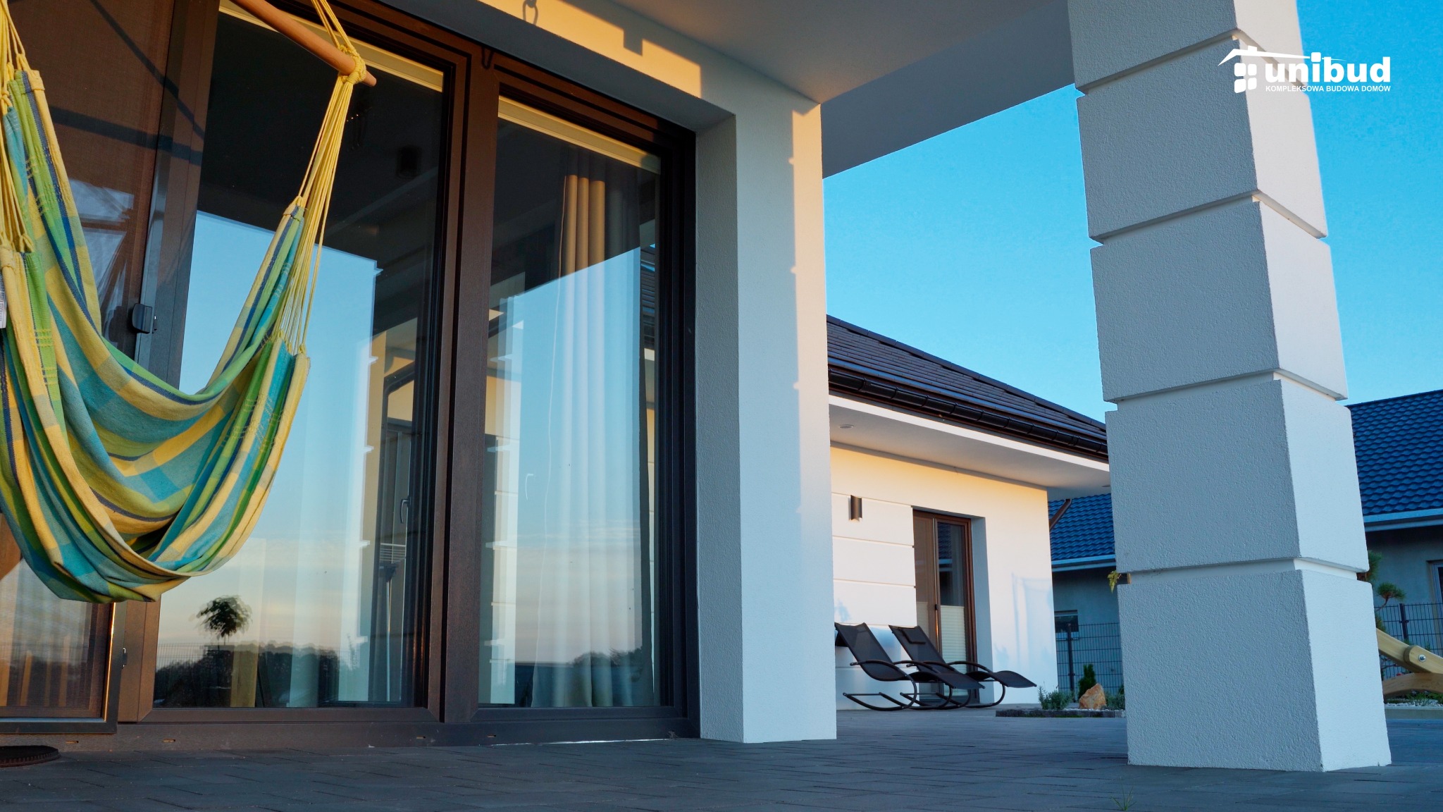 Nowoczesny dom w Miedznej z tarasem, na którym wisi hamak w paski i stoją dwa fotele bujane. Widoczny fragment elewacji z dużymi oknami i kolumną podtrzymującą zadaszenie.