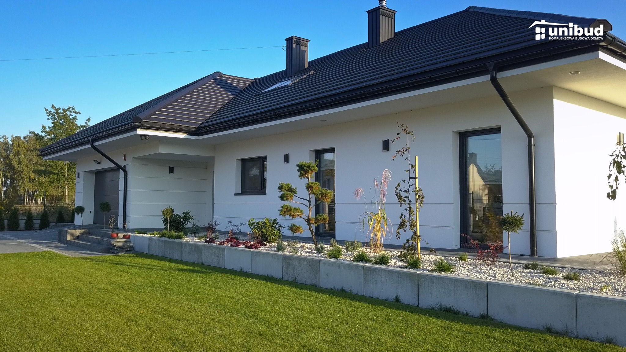 Nowoczesny, parterowy dom z ciemną dachówką i białymi ścianami, ozdobiony minimalistycznym ogrodem z kamieniami i roślinami ozdobnymi, w tle widoczne drzewa.