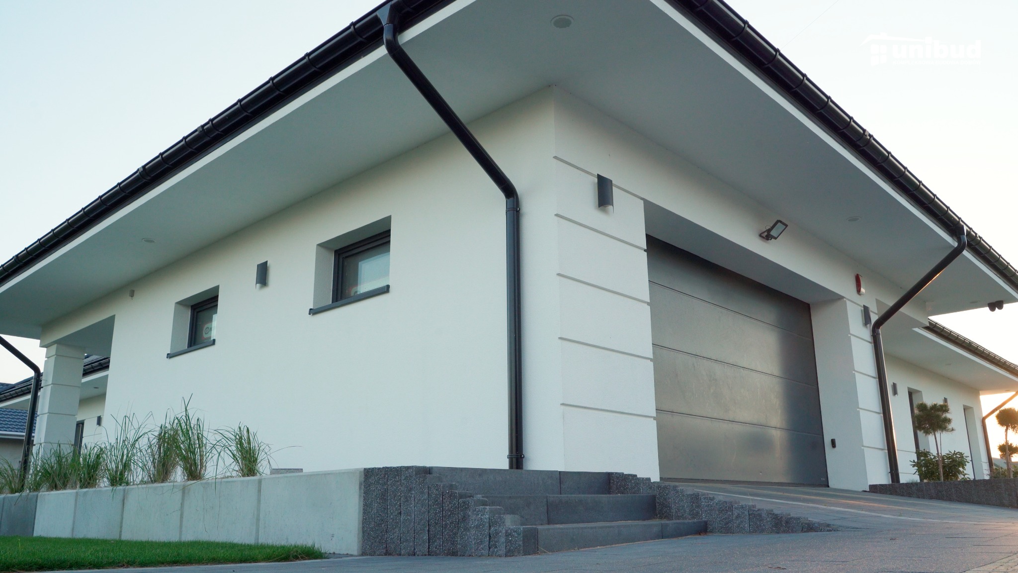 Nowoczesny, parterowy dom z garażem w bryle budynku, wykończony na biało z czarnymi rynnami i grafitową bramą garażową, schody wejściowe wyłożone kamieniem