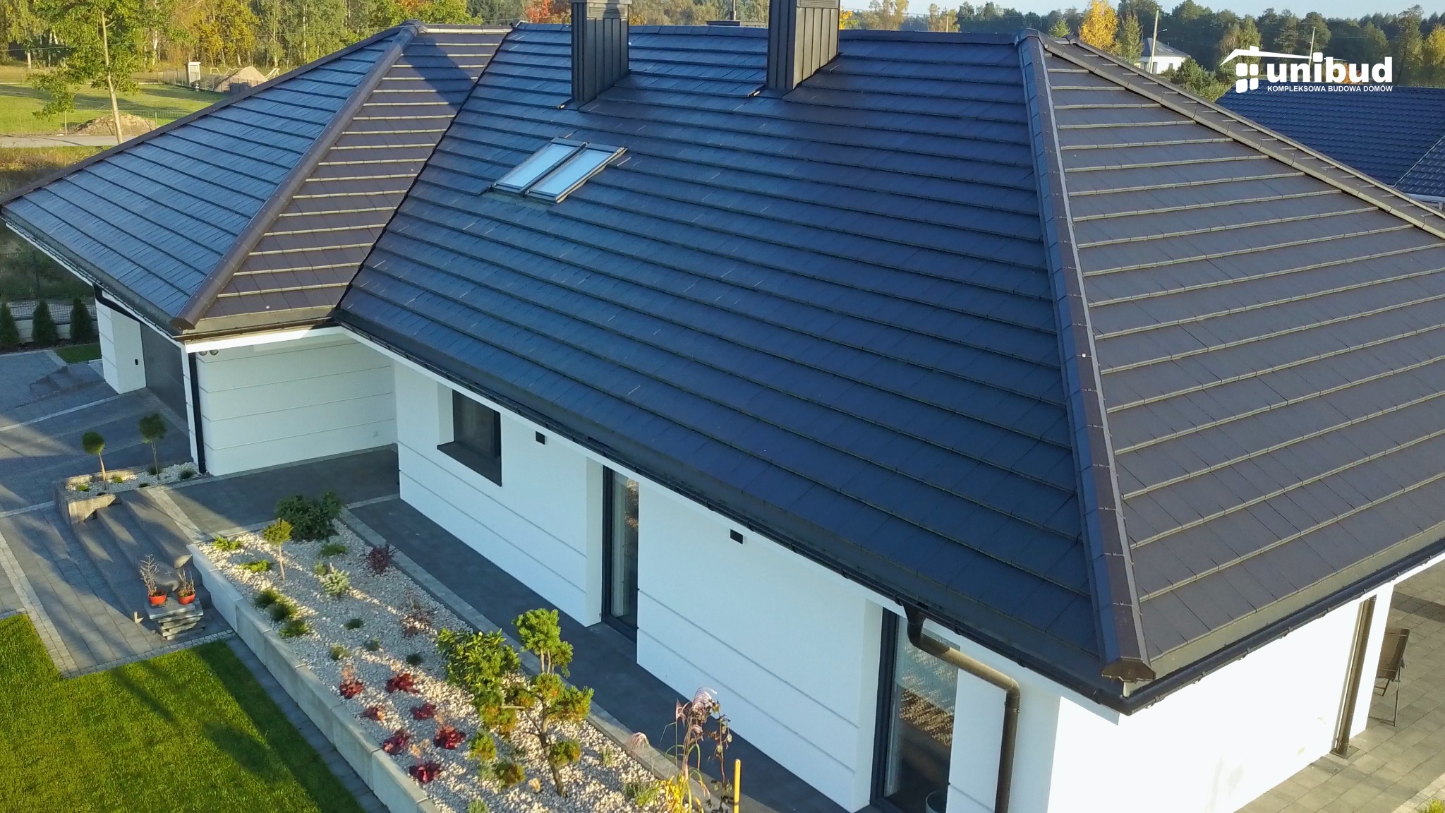 Nowoczesny dom jednorodzinny z ciemną dachówką, dwoma kominami i oknami dachowymi, otoczony zadbanym ogrodem z białym żwirem i niską roślinnością.