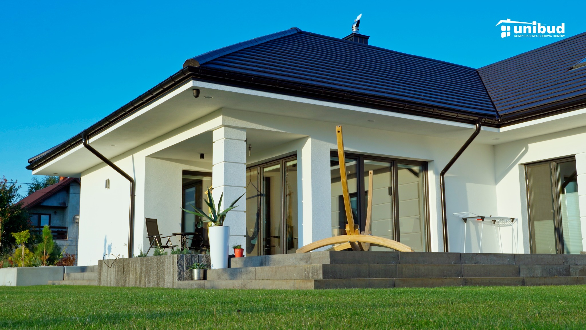 Nowoczesny, parterowy dom z białymi ścianami, ciemnym dachem i dużymi oknami tarasowymi, widziany z poziomu trawnika, z elementami wyposażenia ogrodu na tarasie, pod błękitnym niebem, z logo firmy...