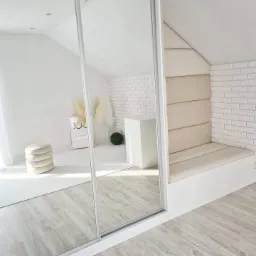 Minimalistyczne wnętrze poddasza z zabudowaną szafą przesuwną z lustrzanymi drzwiami i siedziskiem wnękowym obitym beżową tapicerką, biała cegła na ścianie.
