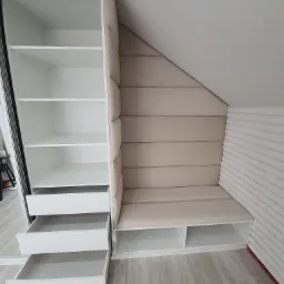 Biała zabudowa pod skosem ze szafą z przesuwnymi drzwiami, tapicerowanym siedziskiem i ścianką, oraz otwartymi szufladami.