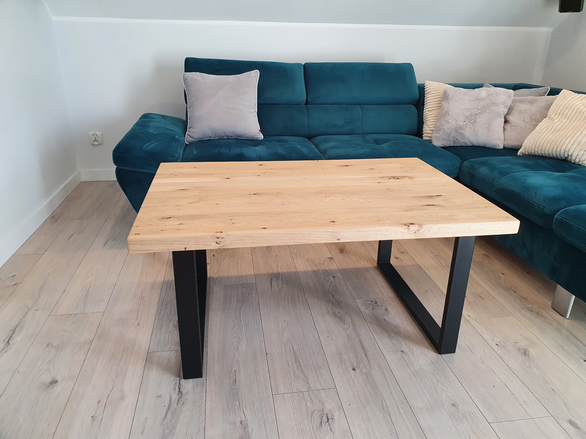 Jasny, drewniany stolik kawowy z czarnymi, metalowymi nogami w kształcie trapezu, ustawiony na panelach podłogowych, z turkusową sofą i poduszkami w tle.