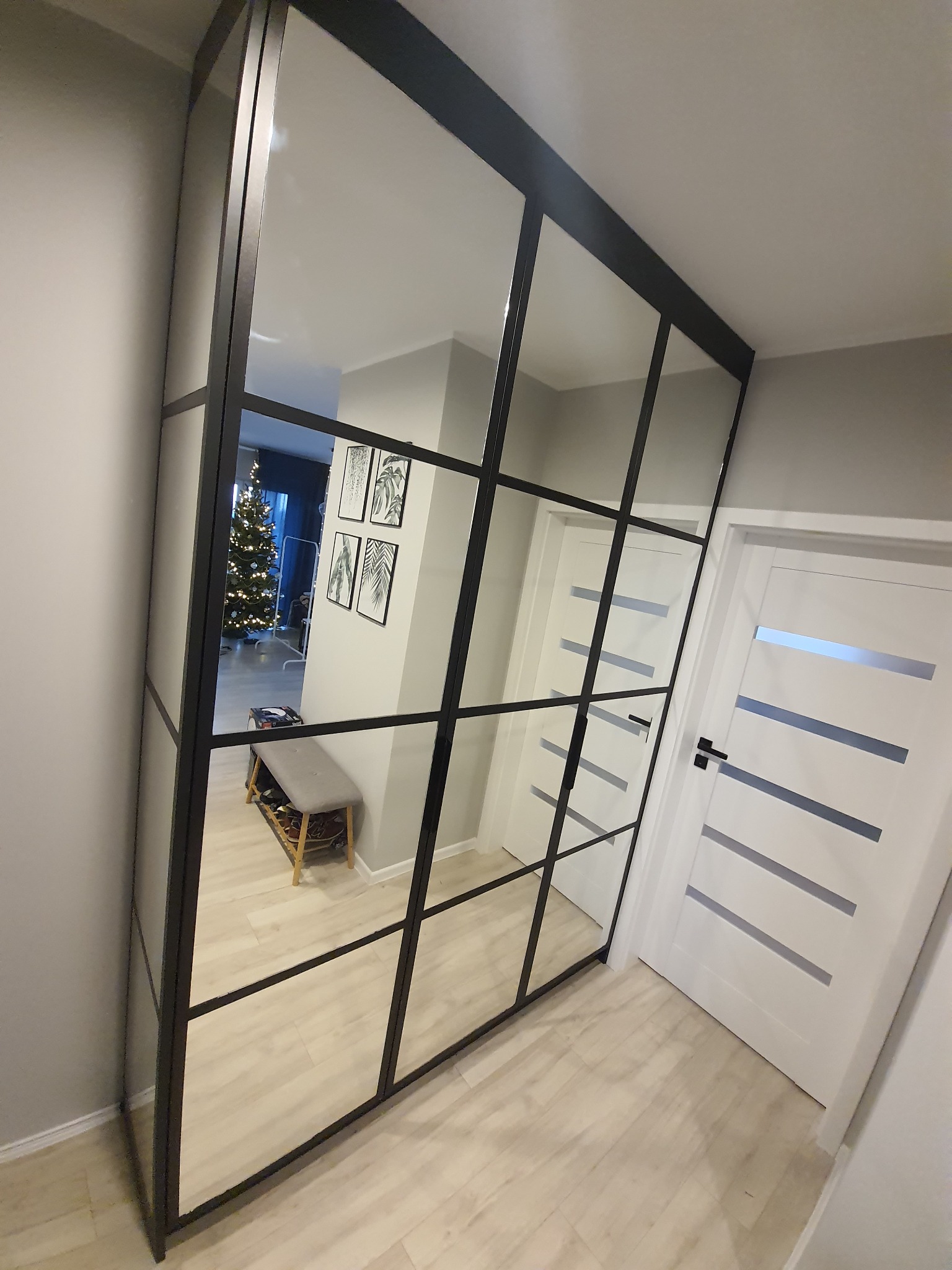 Duża szafa wnękowa z lustrzanymi drzwiami przesuwnymi, czarne metalowe ramy, widoczny fragment przedpokoju z białą ławką i bożonarodzeniową choinką w tle odbicia.