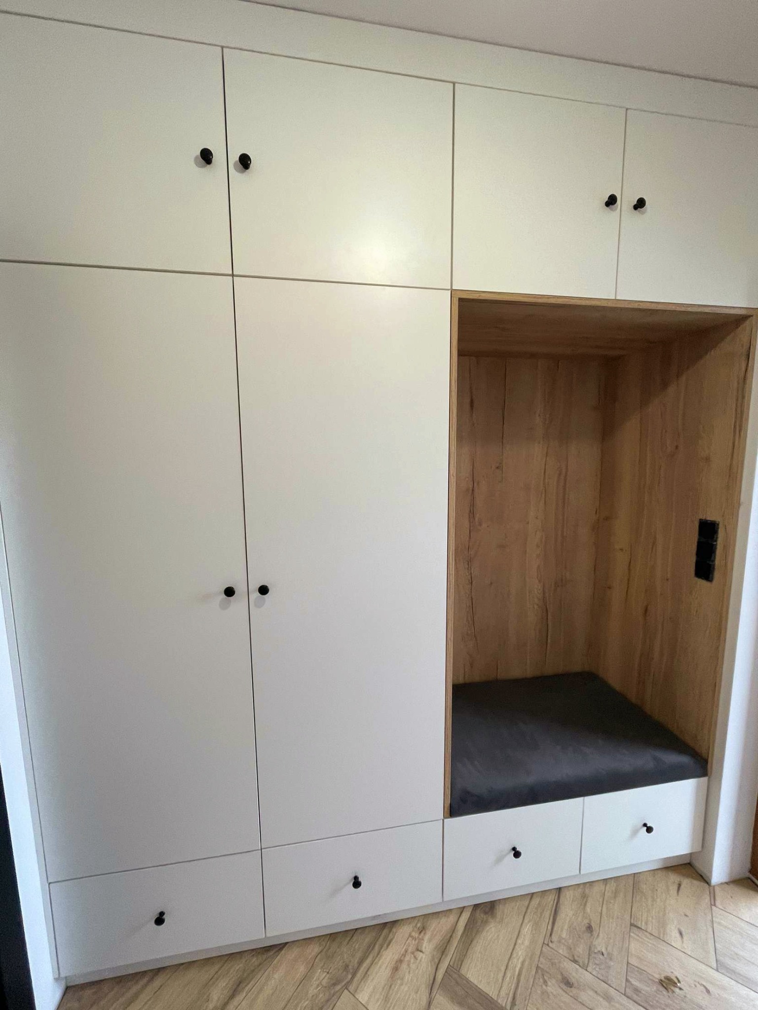 Biała zabudowa meblowa z drewnianą wnęką i czarną poduszką, uzupełniona czarnymi gałkami, na podłodze parkiet ułożony w jodełkę.