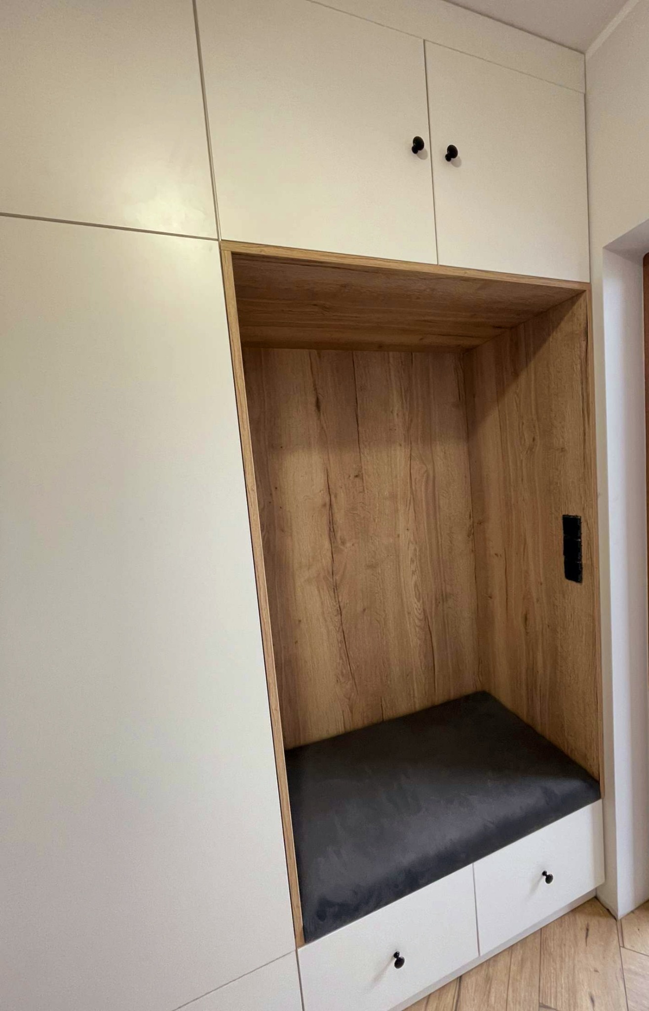 Wnęka z siedziskiem i szufladami w zabudowie meblowej, wykończona drewnopodobną okleiną, z czarnymi uchwytami.