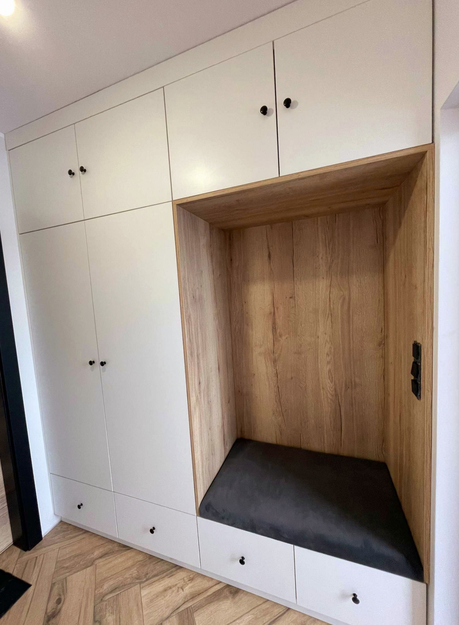 Biała zabudowa meblowa z drewnianą wnęką wykończoną okładziną imitującą drewno, z szarą tapicerowaną ławeczką w środku, na podłodze parkiet w jodełkę.