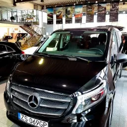 Czarny Mercedes Vito z polską tablicą rejestracyjną w salonie samochodowym, widok z przodu, z widocznymi banerami reklamowymi innych modeli w tle.