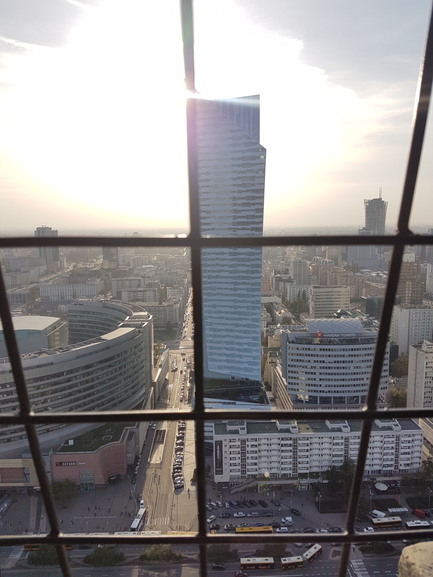 Widok z góry na panoramę miasta z wysokim budynkiem w centrum, widziany przez metalową kratę, z autobusami na ulicach poniżej.