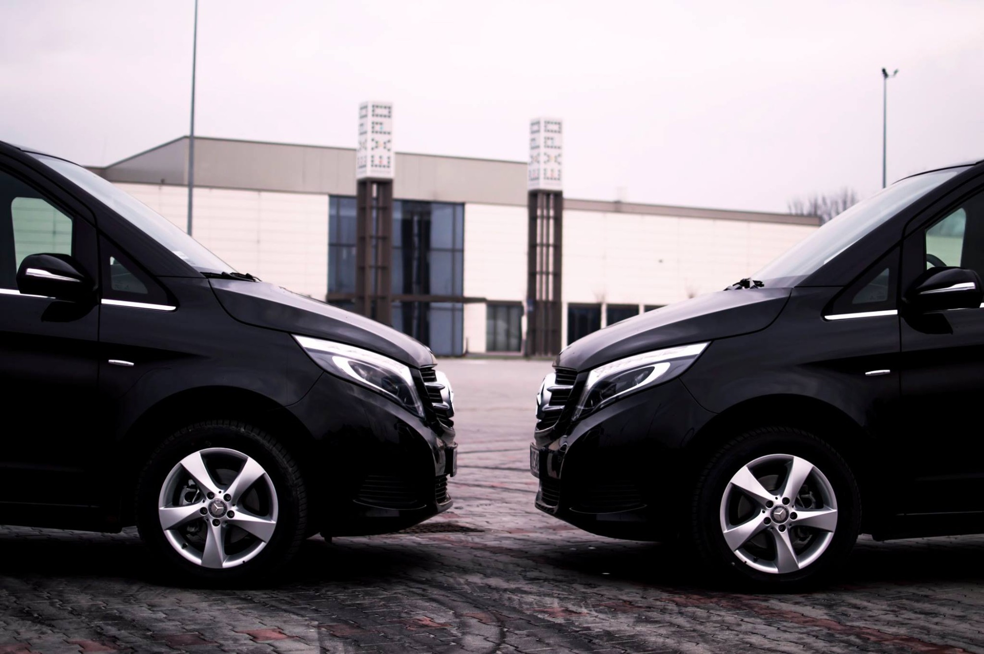 Dwa czarne busy Mercedes V-klasy ustawione przodem do siebie na tle budynku EXPO, z widocznymi srebrnymi felgami.
