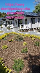 Rząd identycznych, nowoczesnych domków letniskowych z ciemnymi dachami i jasnymi elewacjami, otoczonych starannie zagospodarowanymi rabatami z roślinami iglastymi i korą.