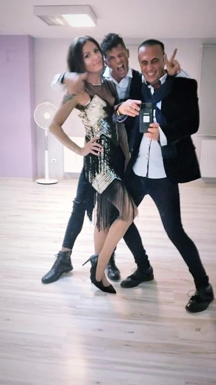 Trzy osoby pozują do zdjęcia w studiu tanecznym: kobieta w cekinowej sukience z frędzlami i butach na obcasie, oraz dwóch mężczyzn w koszulach i marynarkach. Jeden z mężczyzn trzyma telefon i robi...