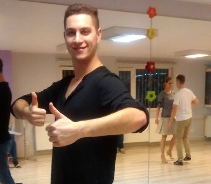 Uśmiechnięty mężczyzna w czarnej koszuli z podniesionymi kciukami na tle sali tanecznej z parą ćwiczącą taniec przed lustrem.