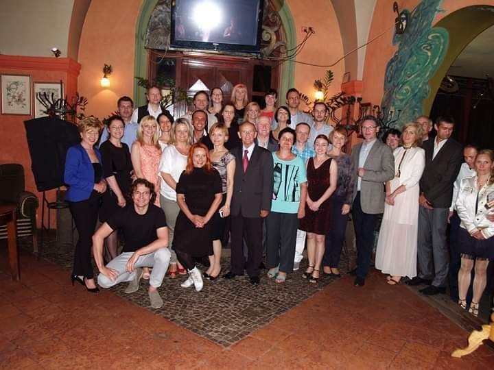 Grupa ludzi pozuje do zdjęcia w sali z dekoracjami, w centrum mężczyzna w czarnym t-shircie kuca.