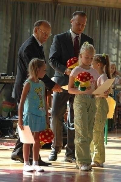 Uroczystość wręczenia nagród młodym tancerkom z pluszowymi grzybami, na scenie dwóch mężczyzn w garniturach i trzy dziewczynki z blond włosami.