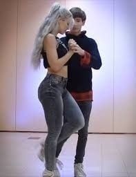 Para tancerzy w trakcie ćwiczeń tanecznych. Kobieta z platynowymi włosami w kucyku, ubrana w czarny top i jeansy, trzyma za rękę mężczyznę w czarnej bluzie. Jasne tło z subtelnym oświetleniem.