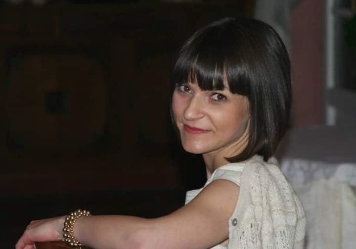 Portret uśmiechniętej kobiety z ciemnymi włosami do ramion, w jasnej bluzce i ażurowym swetrze, siedzącej na krześle i spoglądającej w bok.