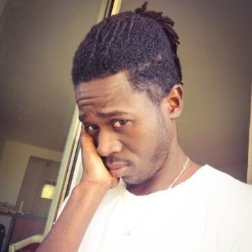 Portret młodego mężczyzny z dredami, ubrany w białą koszulkę, opierającego policzek na dłoni. Wyraz twarzy melancholijny, spojrzenie skierowane w dół. Tło rozmyte, widać fragment drzwi i okna.