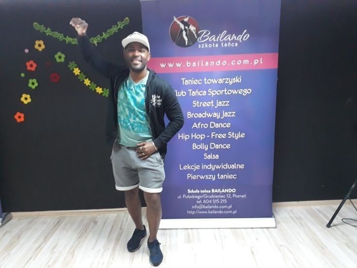 Uśmiechnięty instruktor tańca w sportowym stroju, pozujący na tle ściany z kolorowymi dekoracjami i roll-upu z ofertą szkoły tańca Bailando.
