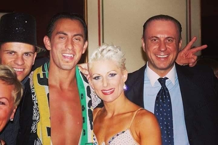 Grupa uśmiechniętych osób, prawdopodobnie tancerzy, pozuje do zdjęcia w Poznaniu, widoczny mężczyzna w garniturze wykonuje gest zwycięstwa.