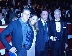 Grupa czterech elegancko ubranych osób, w tym jedna w smokingu, pozuje do zdjęcia na tle widowni w teatrze lub sali koncertowej.