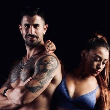 Mężczyzna z tatuażami i łańcuszkami, uśmiechnięty, stoi obok kobiety z długimi włosami i dużymi kolczykami na czarnym tle.