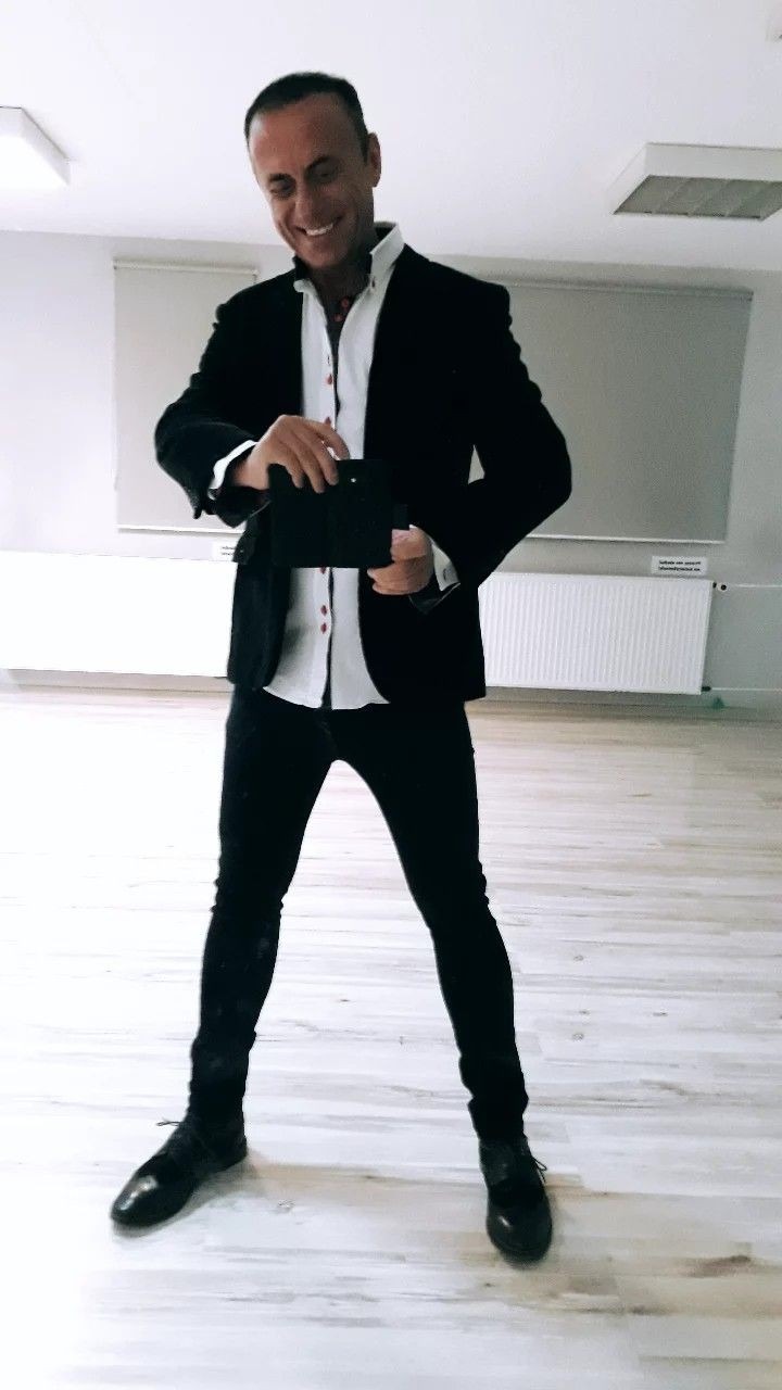 Uśmiechnięty mężczyzna w czarnym garniturze i białej koszuli pozuje w jasnym pomieszczeniu, trzymając w rękach czarny przedmiot przypominający tablet.