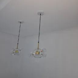 Dwa nowoczesne żyrandole z przezroczystymi, kulistymi kloszami, zawieszone na czarnych przewodach w minimalistycznym wnętrzu z białymi ścianami i sufitami. W rogu widoczny karton z zabawkami.