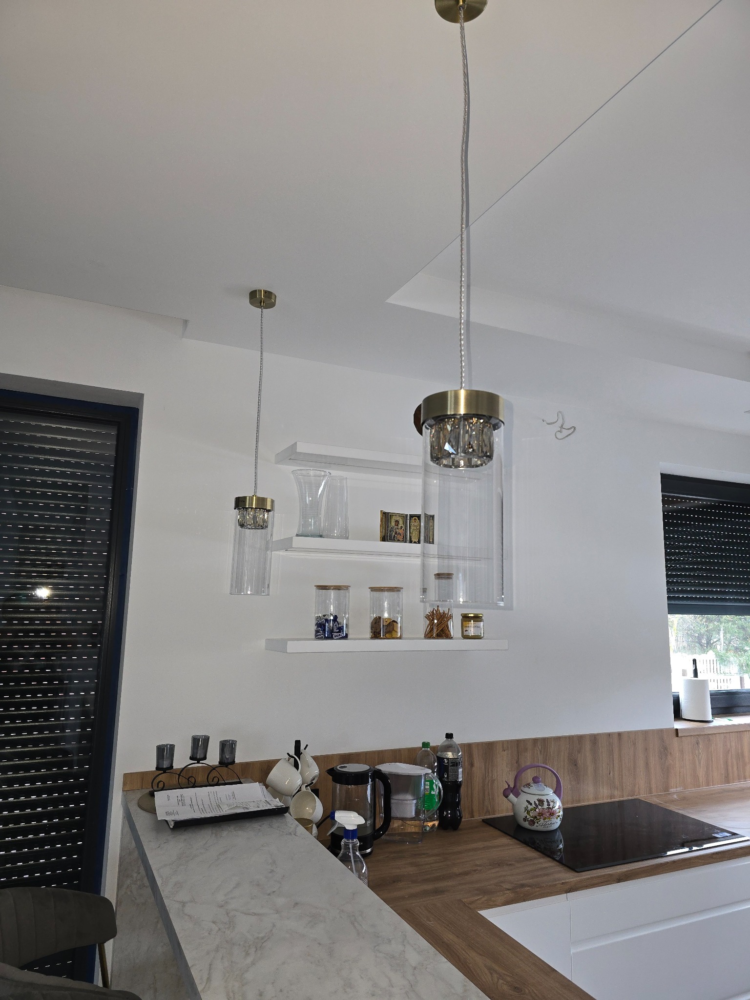 Nowoczesne oświetlenie wiszące z elementami kryształowymi w jasnej kuchni z półkami i roletami okiennymi.