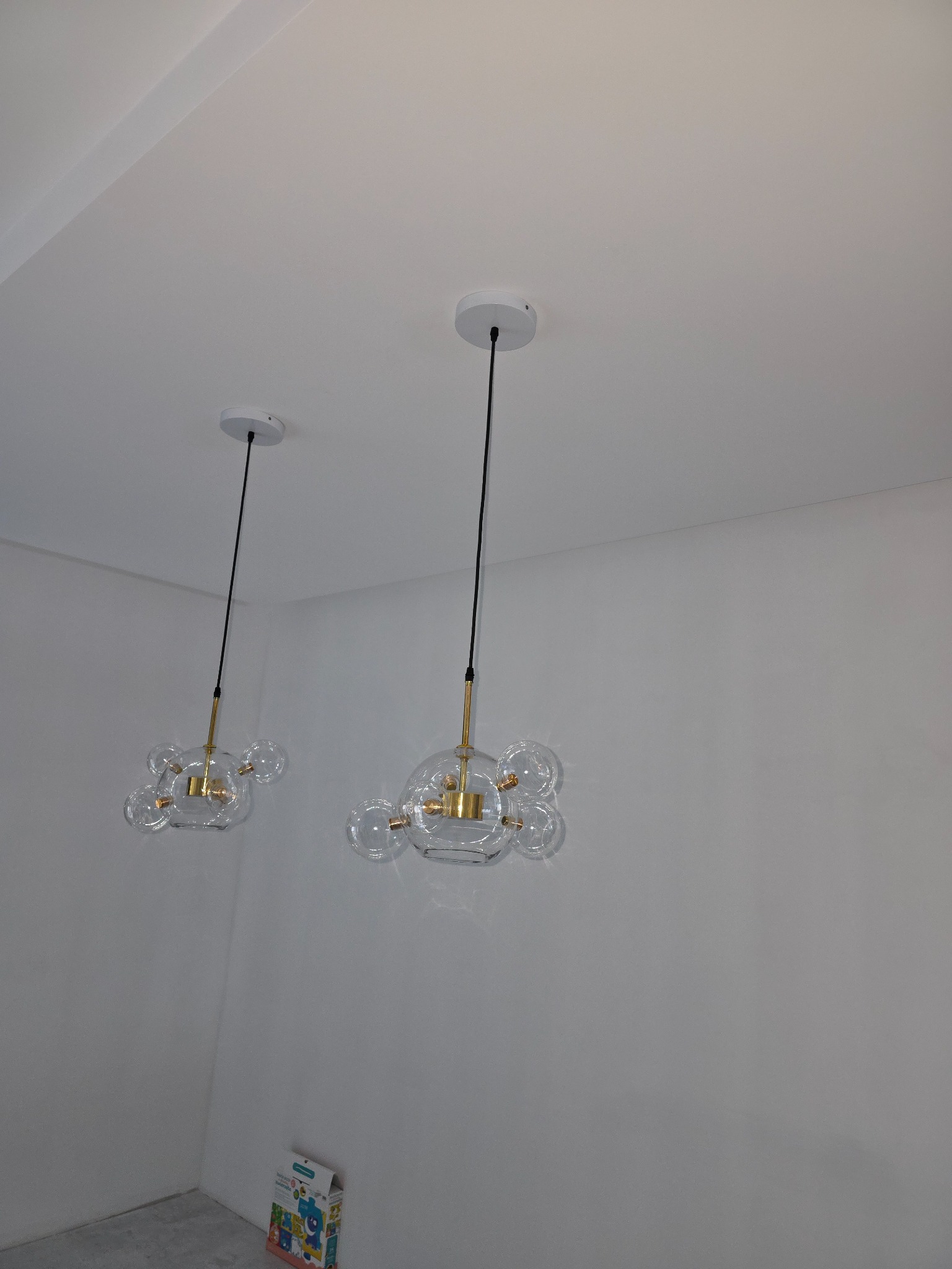 Dwa nowoczesne żyrandole z przezroczystymi, kulistymi kloszami, zawieszone na czarnych przewodach w minimalistycznym wnętrzu z białymi ścianami i sufitami. W rogu widoczny karton z zabawkami.