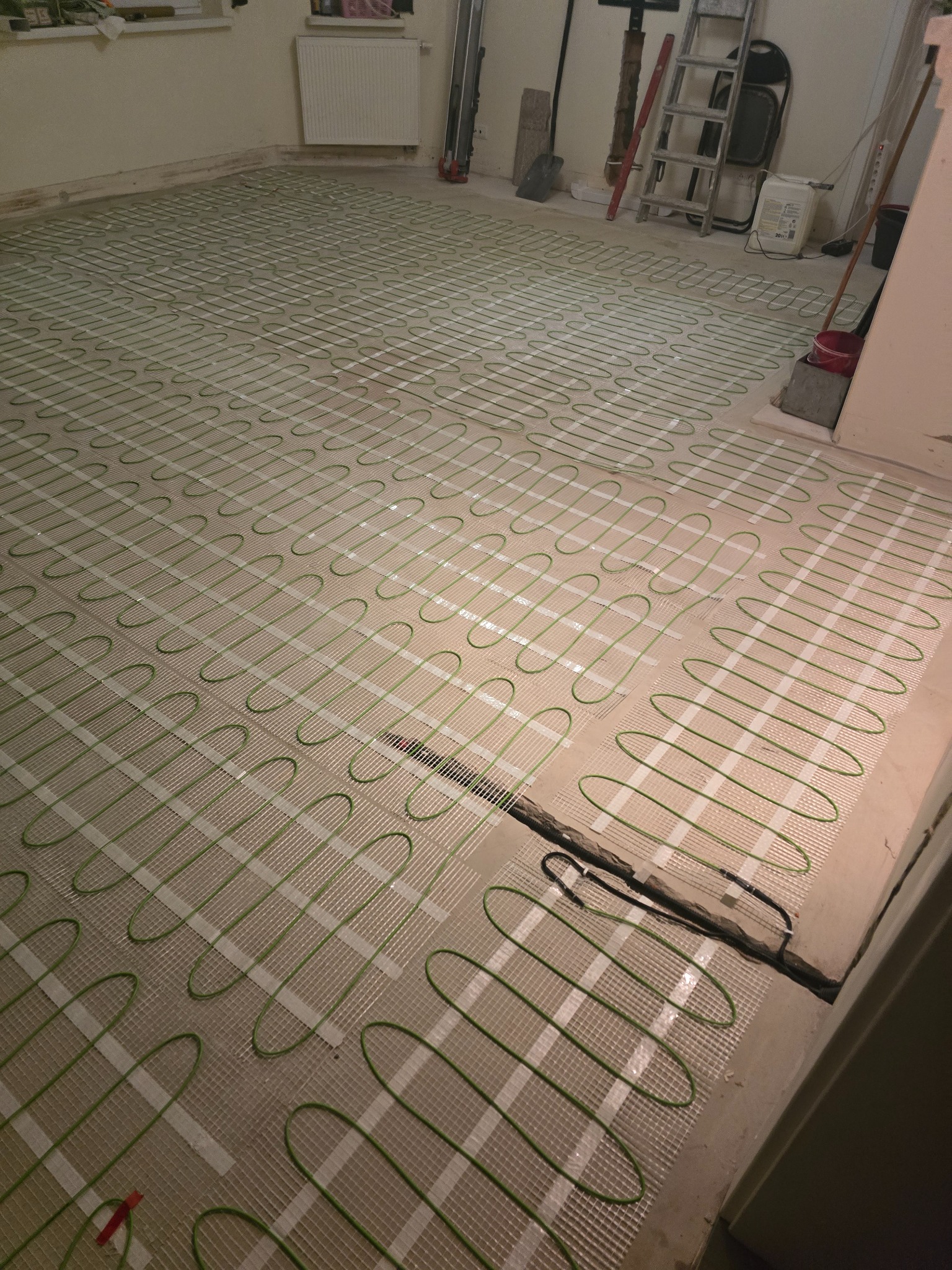 Rozłożona mata grzewcza z zielonymi kablami na podłodze w pomieszczeniu. Widoczne narzędzia i drabina w tle. Etap instalacji ogrzewania podłogowego.