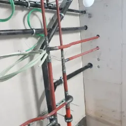 Surowe ujęcie instalacji hydraulicznej i grzewczej w trakcie montażu, widoczne rury miedziane i stalowe, wsporniki, oraz otwory w ścianie z zaznaczonymi wymiarami.