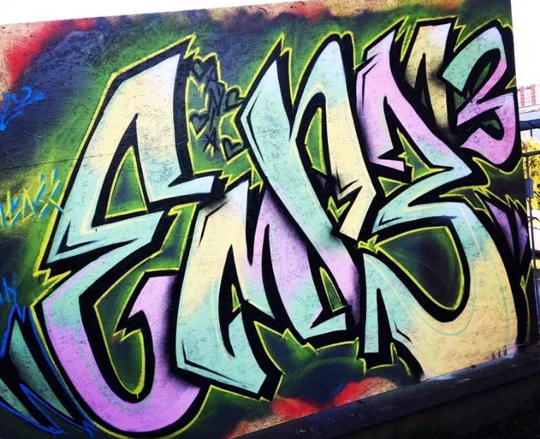 Kolorowe graffiti z dużymi literami na ścianie, wykonane w odcieniach zieleni, różu i żółci, z czarnymi obrysami i elementami dekoracyjnymi.