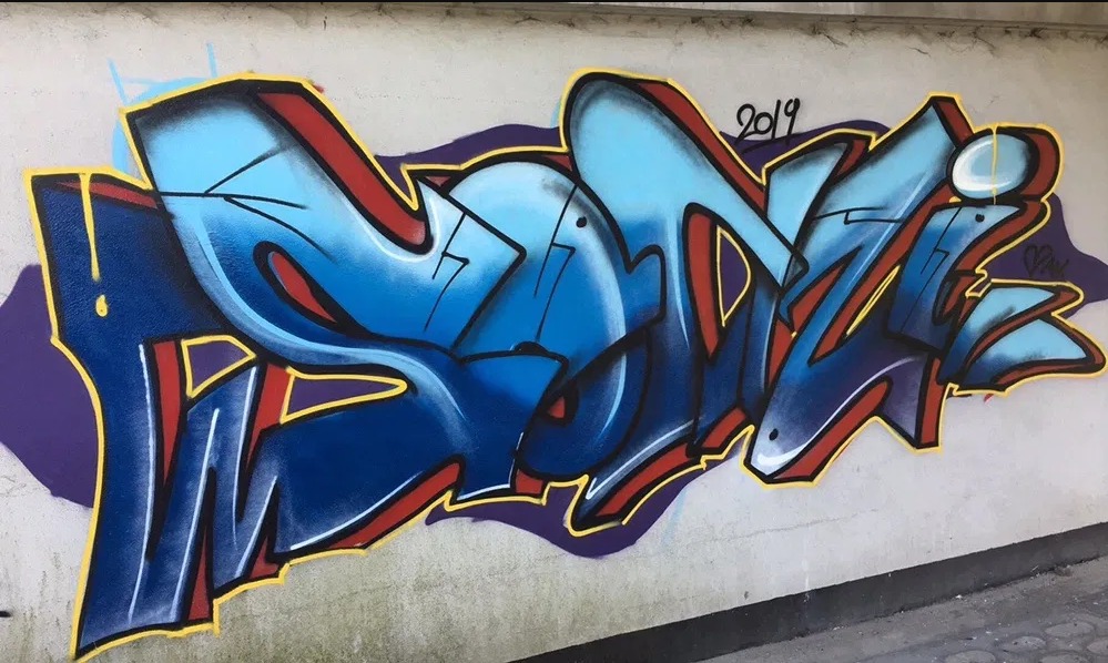 Niebiesko-czerwone graffiti z datą '2019' na szarej ścianie z fioletowym akcentem w tle.