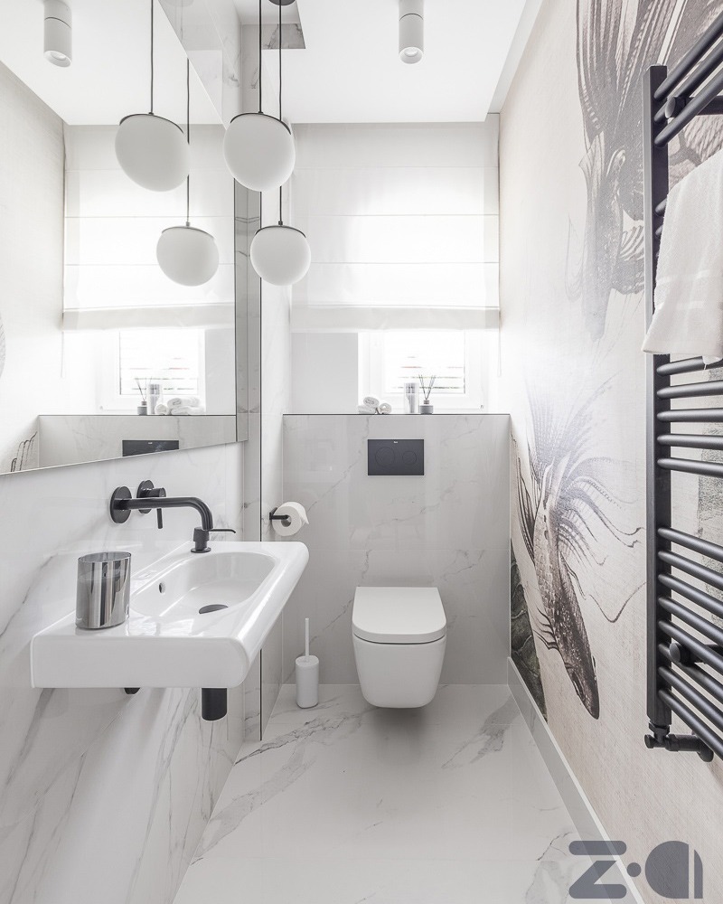 Nowoczesna, wąska łazienka w bieli i szarości, z wiszącą umywalką z czarną armaturą, podwieszaną toaletą i grzejnikiem drabinkowym w kolorze czarnym, akcentowana tapetą z motywem roślinnym...