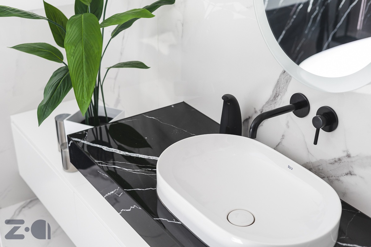 Biała, owalna umywalka nablatowa na czarnym blacie z marmurowym wzorem, czarna bateria ścienna i okrągłe lustro z podświetleniem LED, uzupełnione zieloną rośliną w srebrnej doniczce, na tle białych...