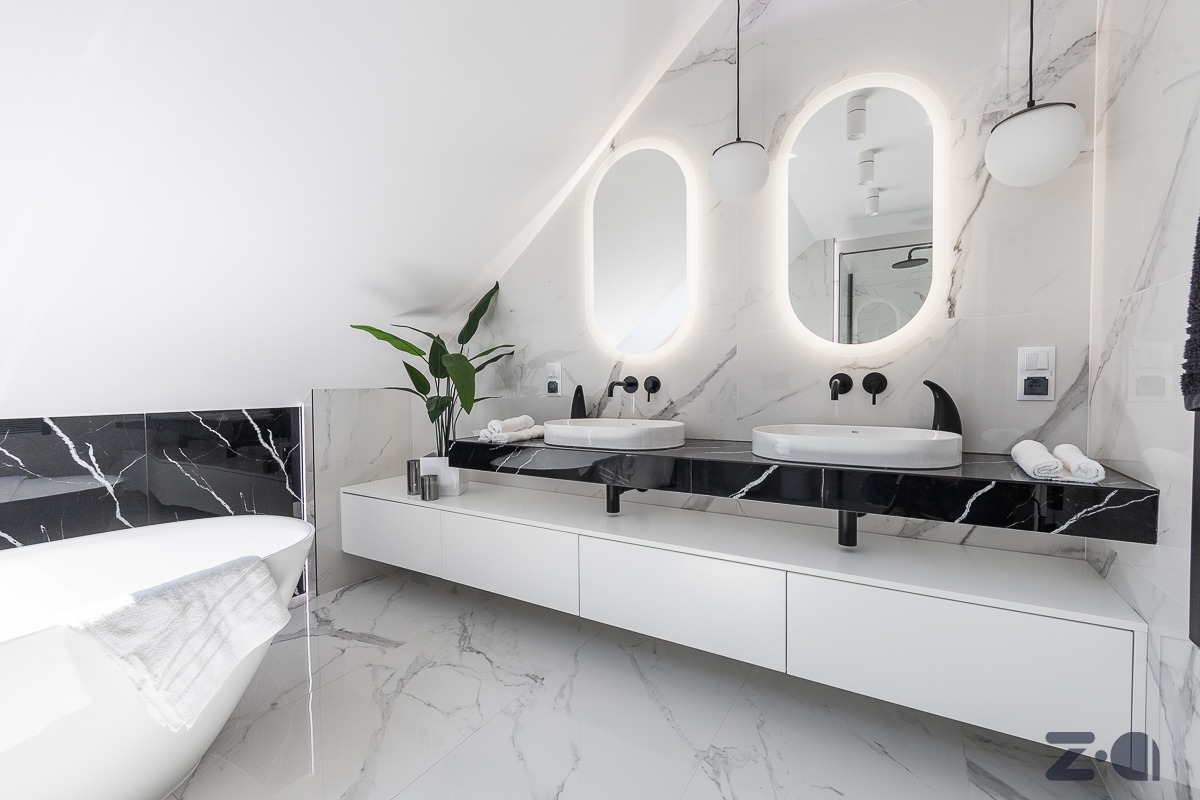 Nowoczesna łazienka na poddaszu z marmurową podłogą i ścianami, owalnymi lustrami z podświetleniem LED, czarnym blatem z dwoma białymi umywalkami, wolnostojącą wanną i zieloną rośliną w doniczce.