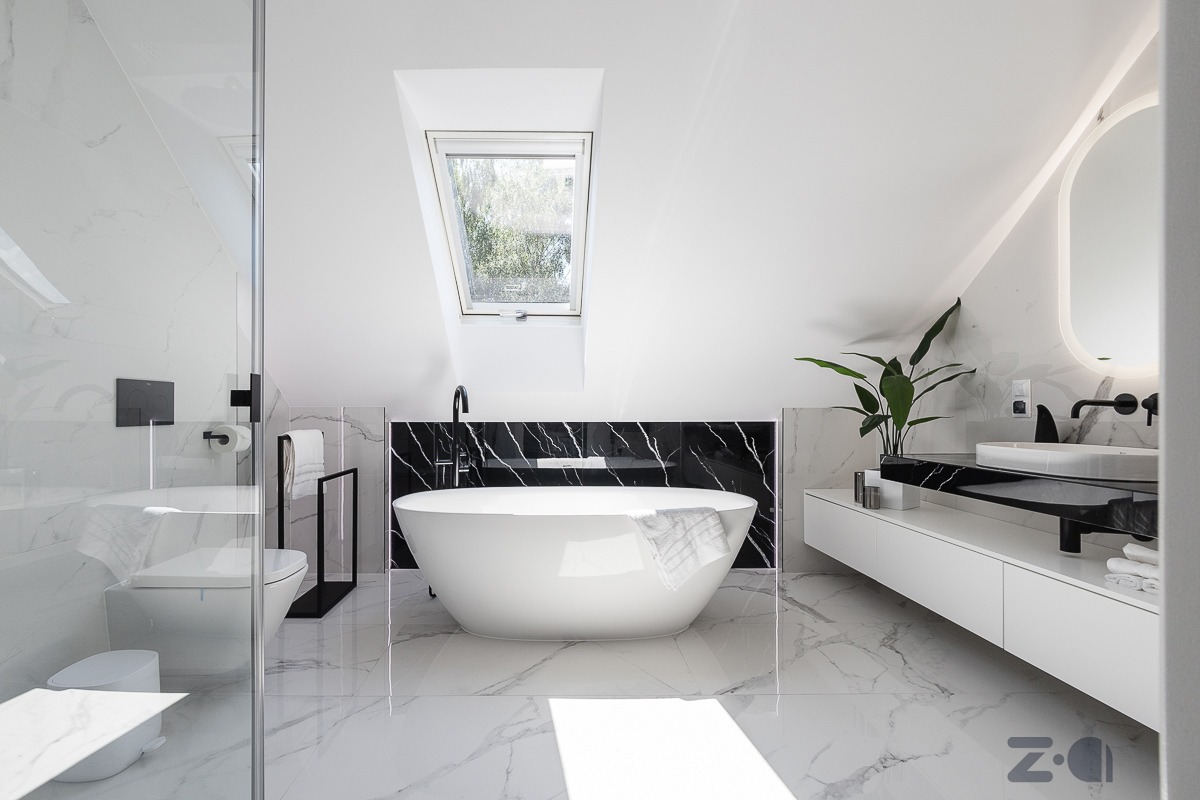 Nowoczesna łazienka na poddaszu z białą, wolnostojącą wanną na tle czarnej ściany z marmurowym wzorem, białą umywalką na szafce i kabiną prysznicową w minimalistycznym stylu.