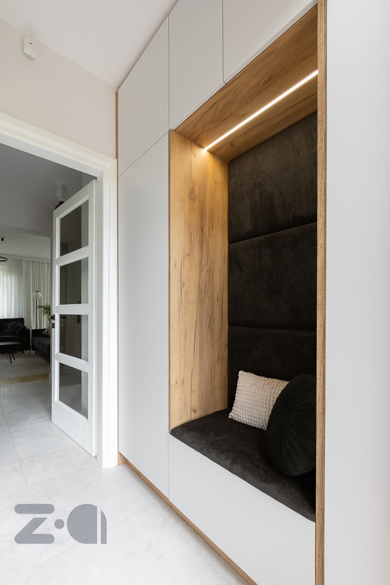 Wnęka wypoczynkowa z podświetleniem LED w przedpokoju, obita ciemnoszarym welurem, z jasną kwadratową i ciemną okrągłą poduszką, obudowana białymi szafkami i okładziną drewnopodobną.