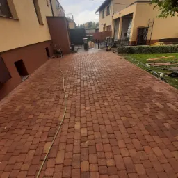 Nowo ułożona, czerwono-brązowa kostka brukowa na podjeździe między budynkami, widoczny wąż ogrodowy, w tle brama wjazdowa i narzędzia.
