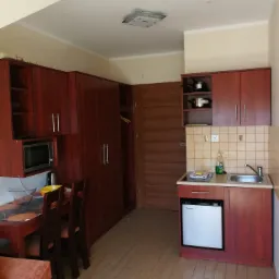 Wnętrze pokoju hotelowego z aneksem kuchennym, widoczny stół z krzesłami, szafa, zabudowa kuchenna z mikrofalówką i lodówką.