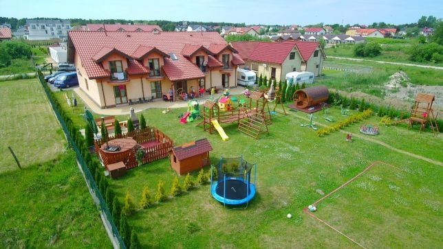 Widok z góry na ośrodek wypoczynkowy z placem zabaw, domkami z czerwonymi dachami i zielonym trawnikiem, w tle zabudowa mieszkalna.