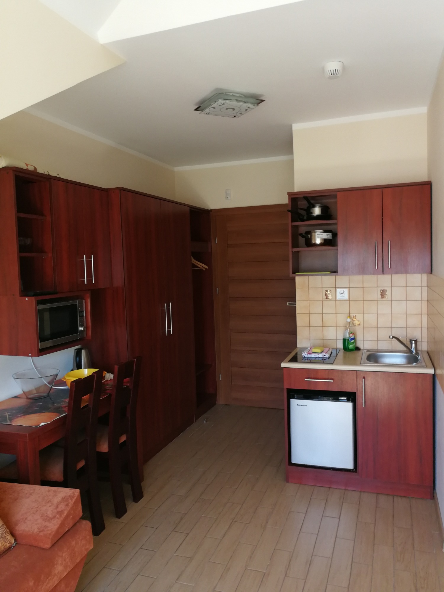 Wnętrze pokoju hotelowego z aneksem kuchennym, widoczny stół z krzesłami, szafa, zabudowa kuchenna z mikrofalówką i lodówką.