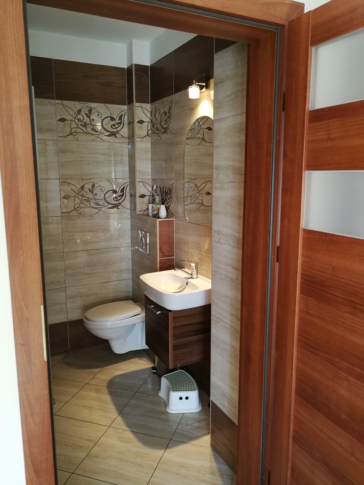 Nowoczesna łazienka z beżowymi płytkami zdobionymi motywami roślinnymi, białą umywalką z szafką w kolorze ciemnego drewna, toaletą i małym białym stołeczkiem dla dzieci, widziana zza drewnianej...