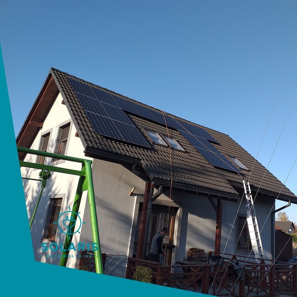 Dom jednorodzinny z zamontowanymi panelami słonecznymi na dachu, drabina oparta o budynek, widoczne okna dachowe i błękitne niebo w tle. Częściowo zasłonięte logo firmy w lewym dolnym rogu.