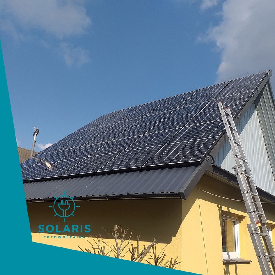 Żółty dom z szarym dachem, na którym zamontowane są panele słoneczne; aluminiowa drabina oparta o dach, błękitne niebo w tle.