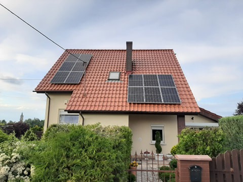 Dom jednorodzinny z czerwoną dachówką i panelami słonecznymi na dachu, widok z poziomu ulicy w Kożuchowie, z widoczną wieżą kościoła w tle.