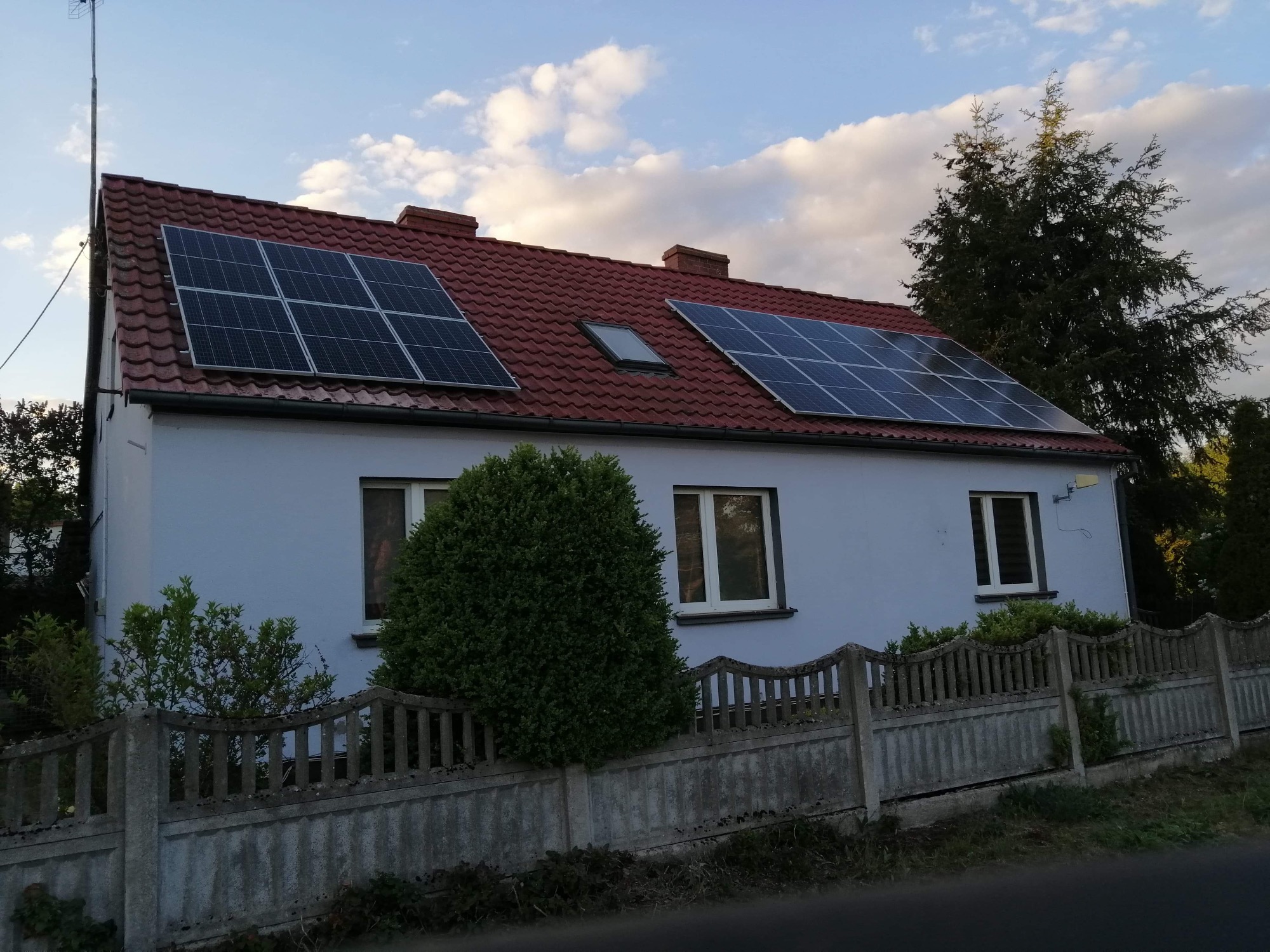 Dom jednorodzinny z czerwonym dachem i dwoma zestawami paneli słonecznych, widoczne zza betonowego ogrodzenia w Chrośnicy.